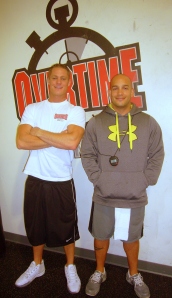 Matt Larkin and Jorge Aguirre, certified fitness trainers, post in front of Overtime Fitness gym wall logo